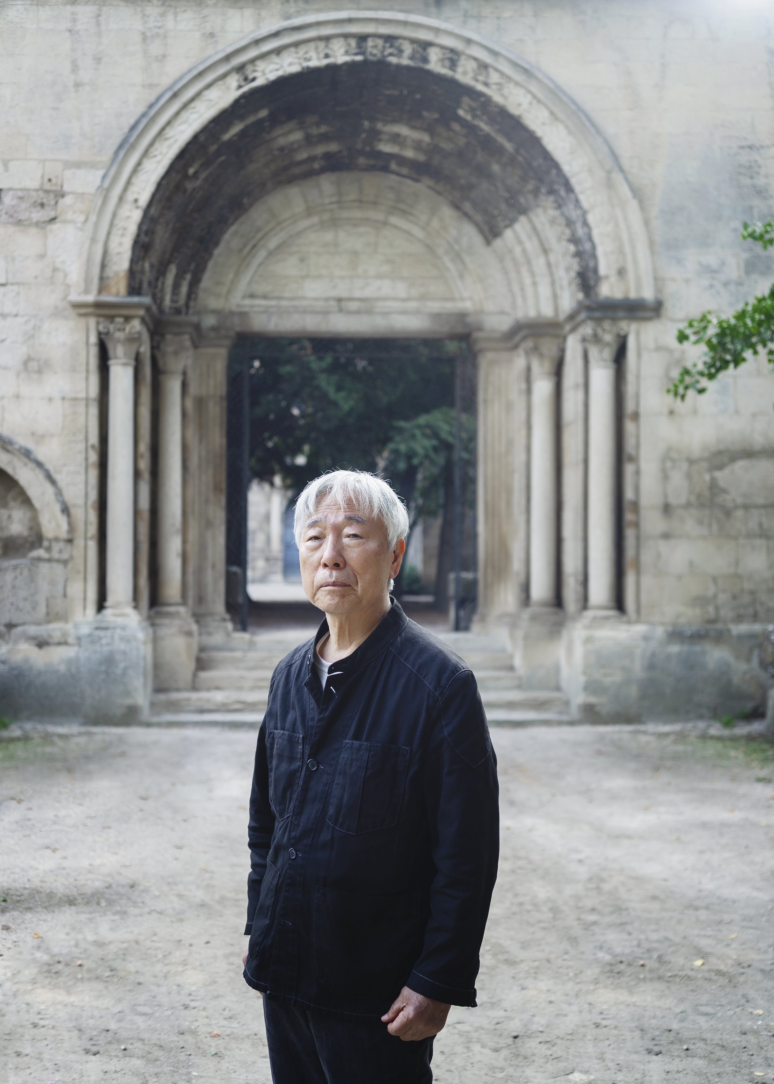 Lee Ufan, Foto: Claire Dorn, Courtesy Studio Lee Ufan Ein älterer Mann steht in schwarzer Kleidung vor einem Toreingang aus Steinen. Der Mann schaut ernst und steht leicht abgewandt.