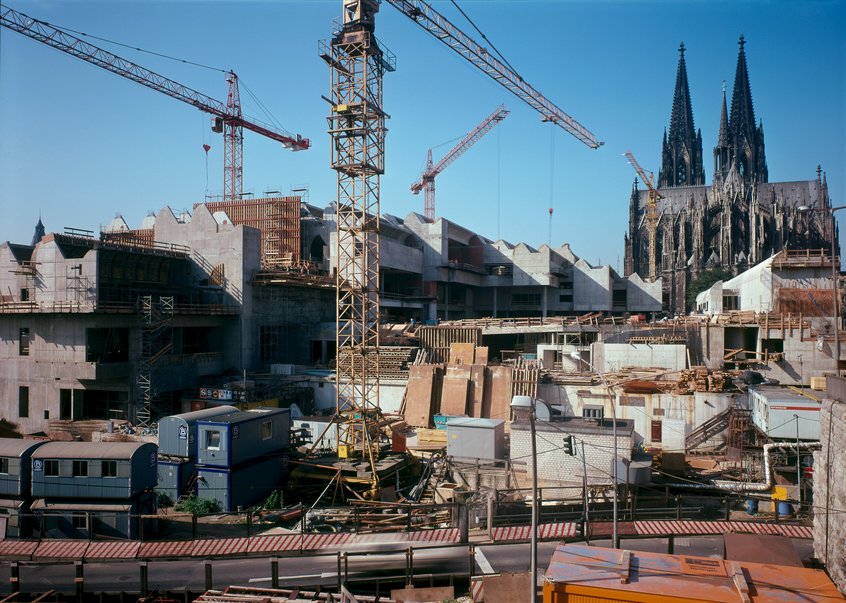 Farbfotografie einer großen innerstädtischen Baustelle. Mehrere Baukräne ragen über unfertige Gebäudeteile mit markanten, gezackten Dachformen. Im Vordergrund stehen Baucontainer, Absperrungen, Baumaterialien und Baustelleneinrichtungen. Im Hintergrund ist der Kölner Dom mit seinen beiden hohen Türmen zu sehen. Über der Baustelle spannt sich ein klarer blauer Himmel.