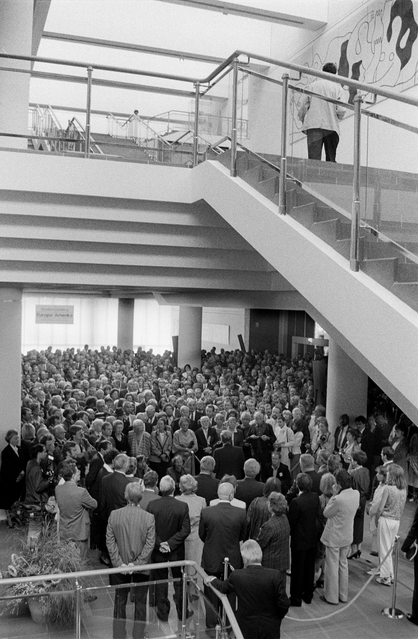 Schwarz-Weiß-Fotografie einer dicht gedrängten Menschenmenge in einem mehrstöckigen Gebäude. Im Vordergrund und im oberen Bildbereich verlaufen Treppen und Geländer. Viele Menschen stehen im Eingangs- oder Ausstellungsbereich und blicken in Richtung Bildmitte. Auf der oberen Etage steht eine Person am Geländer. Die Aufnahme vermittelt den Eindruck einer stark besuchten Veranstaltung.