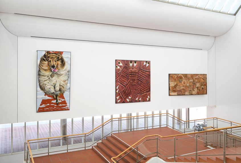 Jana Euler, Triptych – Image Viewer Connections, 2026, Schultze Projects #5, Museum Ludwig, Köln © Jana Euler, Foto: Historisches Archiv der Stadt Köln mit Rheinischem Bildarchiv//Marc Weber Blick auf ein helles Treppenhaus mit zwei symmetrisch angeordneten Treppen und Holzhandläufen, die zu einem Zwischenpodest führen. An der weißen Wand drüber hängen drei großformatige Kunstwerke: links ein Gemälde eines Langhaar Collies, der auf die Betrachter*innen zuzulaufen scheint. In der Mitte ein abstraktes Bild in Rot- und Brauntönen mit geometrischen Mustern und Augen, die einen ansehen. Rechts ein abstraktes Werk in Beige- und Brauntönen mit kreisförmigen und organischen Formen.