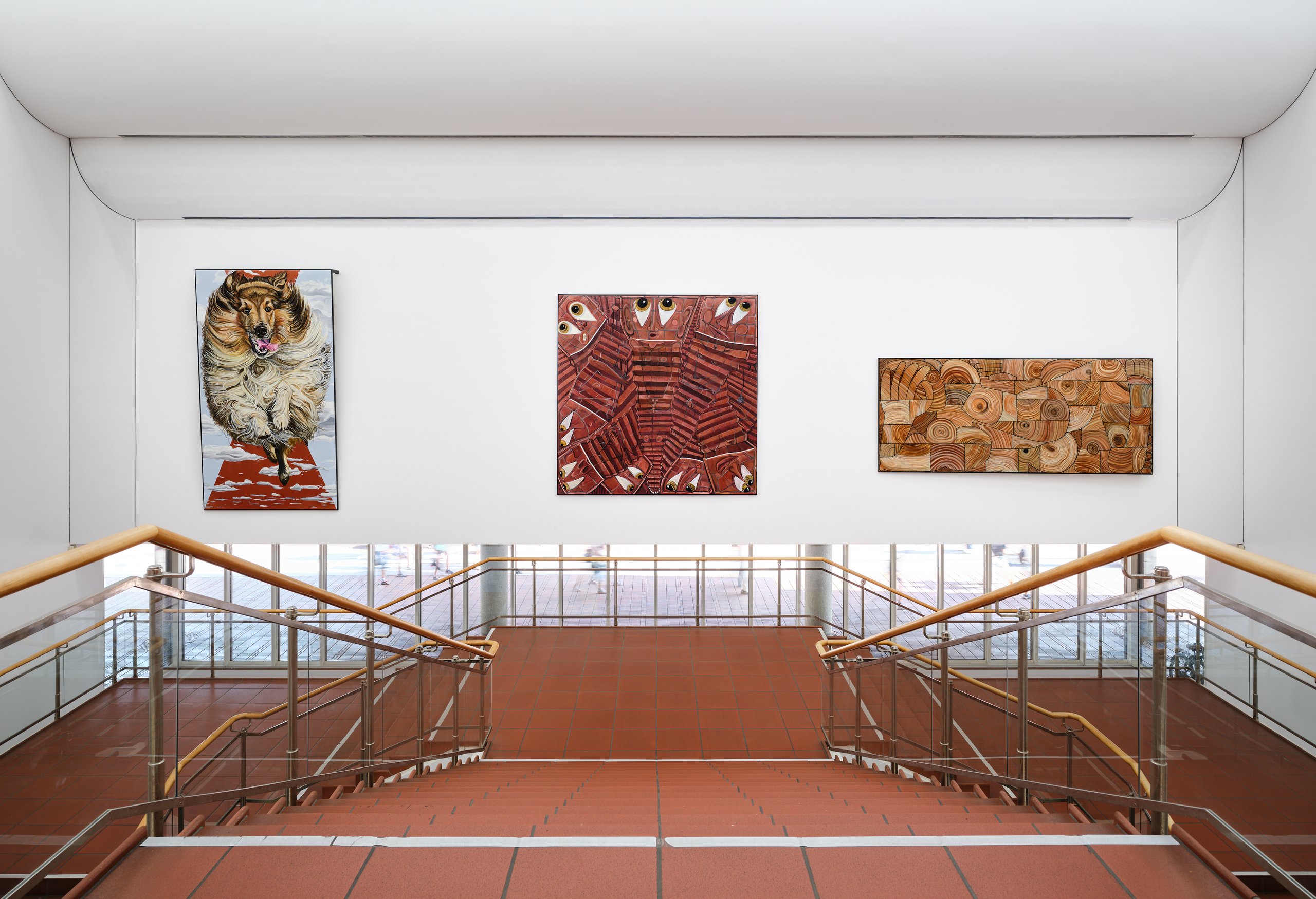 Jana Euler, Triptych – Image Viewer Connections, 2026, Schultze Projects #5, Museum Ludwig, Köln © Jana Euler, Foto: Historisches Archiv der Stadt Köln mit Rheinischem Bildarchiv//Marc Weber Blick auf ein helles Treppenhaus mit zwei symmetrisch angeordneten Treppen und Holzhandläufen, die zu einem Zwischenpodest führen. An der weißen Wand drüber hängen drei großformatige Kunstwerke: links ein Gemälde eines Langhaar Collies, der auf die Betrachter*innen zuzulaufen scheint. In der Mitte ein abstraktes Bild in Rot-und Brauntönen mit geometrischen Mustern und Augen, die einen ansehen. Rechts ein abstraktes Werk in Beige- und Brauntönen mit kreisförmigen und organischen Formen.