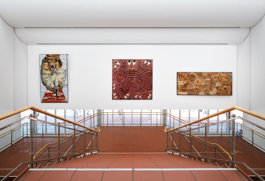 Jana Euler, Triptych – Image Viewer Connections, 2026, Schultze Projects #5, Museum Ludwig, Köln © Jana Euler, Foto: Historisches Archiv der Stadt Köln mit Rheinischem Bildarchiv//Marc Weber Blick auf ein helles Treppenhaus mit zwei symmetrisch angeordneten Treppen und Holzhandläufen, die zu einem Zwischenpodest führen. An der weißen Wand drüber hängen drei großformatige Kunstwerke: links ein Gemälde eines Langhaar Collies, der auf die Betrachter*innen zuzulaufen scheint. In der Mitte ein abstraktes Bild in Rot-und Brauntönen mit geometrischen Mustern und Augen, die einen ansehen. Rechts ein abstraktes Werk in Beige- und Brauntönen mit kreisförmigen und organischen Formen.