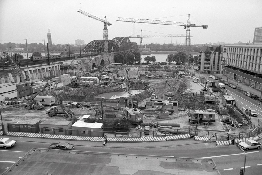 Schwarz-Weiß-Aufnahme einer großflächigen Baustelle in einem Stadtgebiet. Im Vordergrund stehen Baucontainer, Fahrzeuge und Baumaschinen. Dahinter sind zahlreiche Baugruben, Gerüste und Baustellenmaterialien zu sehen. Im Hintergrund erhebt sich eine große Bogenbrücke über einen Fluss oder ein Gewässer. Mehrere Baukräne ragen über das Gelände, und links ist ein Zug zu sehen. Im Hintergrund erstreckt sich die städtische Bebauung, so wie eine Brücke.