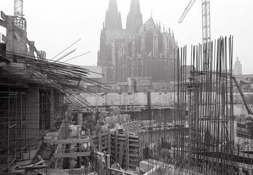 Schwarz-weiß Fotografie einer großen Baustelle. Im Hintergrund steht ein imposanter gotischer Kölner Dom mit zwei hohen Türmen. Im Vordergrund sind zahlreiche Baustellenkonstruktionen, Holzbretter, Betonwände und dicht stehende Stahlbewehrungen zu sehen. Mehrere Baukräne ragen in den Himmel. Die Aufnahme vermittelt den Eindruck einer historischen Stadt während einer umfangreichen Bauphase.