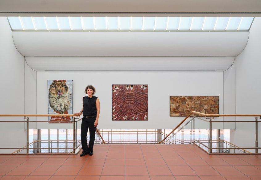 Jana Euler vor ihrer Arbeit Triptych – Image Viewer Connections, 2026 Schultze Projects #5, Museum Ludwig, Köln © Jana Euler Foto: Historisches Archiv der Stadt Köln mit Rheinischem Bildarchiv//Marc Weber Blick auf ein helles Treppenhaus mit zwei symmetrisch angeordneten Treppen und Holzhandläufen, die zu einem Zwischenpodest führen. An der weißen Wand drüber hängen drei großformatige Kunstwerke: links ein Gemälde eines Langhaar Collies, der auf die Betrachter*innen zuzulaufen scheint. In der Mitte ein abstraktes Bild in Rot- und Brauntönen mit geometrischen Mustern und Augen, die einen ansehen. Rechts ein abstraktes Werk in Beige- und Brauntönen mit kreisförmigen und organischen Formen. Im Vordergrund steht eine in schwarz gekleidete Person